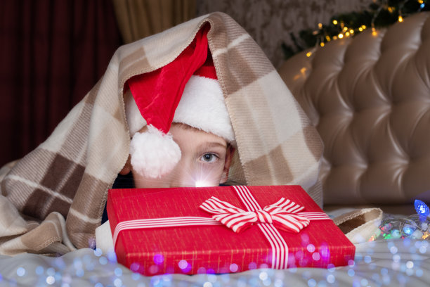 一个可爱的白人男孩戴着圣诞帽，藏在被窝里打开圣诞老人送的礼物图片下载