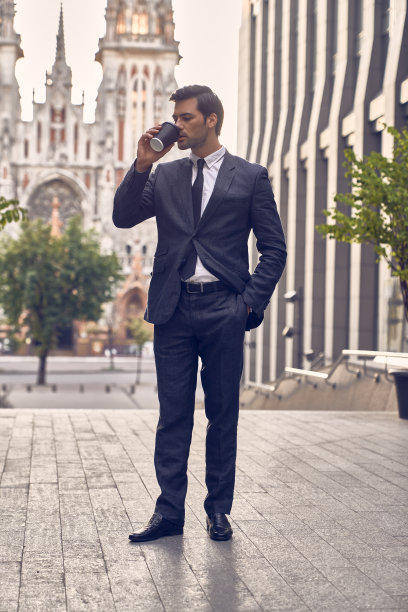 咖啡休息。自信的年轻人，穿着全套西装，拿着咖啡杯，看着别处，站在户外，以城市景观为背景。图片下载