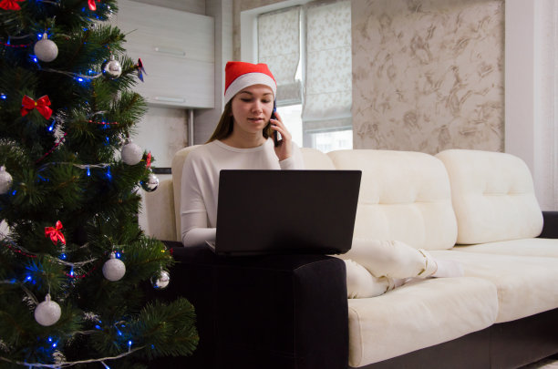 一个戴着医用口罩的女孩在圣诞树后面的笔记本电脑后面的手机上写了一条信息。圣诞节在网上工作的女孩图片下载