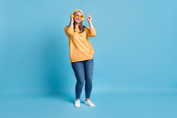 可爱迷人的老妇人的全身大小的照片听音乐拿着耳机直接手指跳舞独自在家哼歌戴太阳镜黄色套头衫鲜艳的蓝色背景图片下载