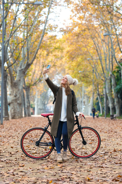 美丽的年轻女子与她的智能手机自拍时，通过公园在秋天。图片下载