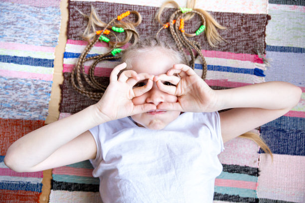 特写照片中，一个梳着辫子的快乐女孩躺在编织的条纹地毯上，微笑着，交叉着手指戴着眼镜。图片下载