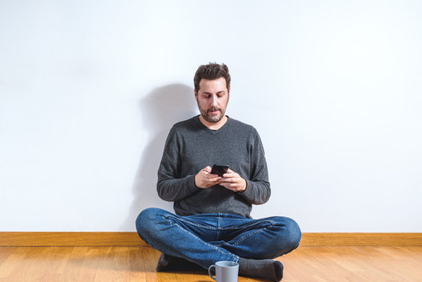 一个40多岁的年轻人坐在空荡荡的公寓地板上玩着智能手机图片下载