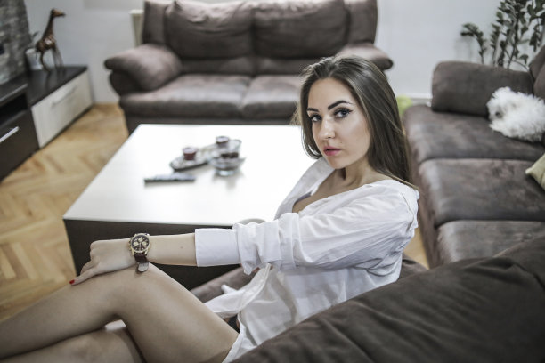 一个穿着男士衬衫的漂亮年轻女子正坐在她的家里，坐在客厅的沙发上，享受着性爱和一个自由的周末。图片下载