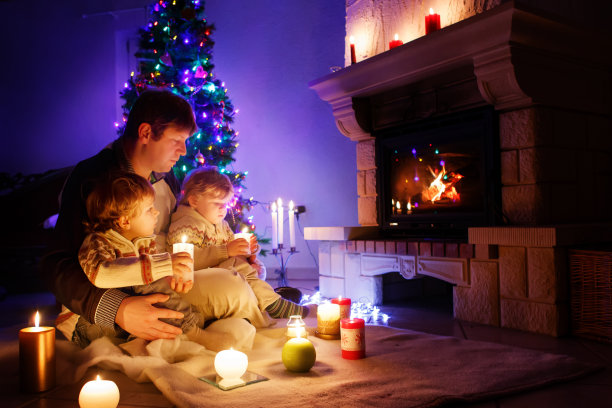 父亲和两个蹒跚学步的小男孩坐在烟囱、蜡烛和壁炉旁，看着火苗。家庭庆祝圣诞节。背景是圣诞树和彩灯。孩子们喜欢礼物素材图片