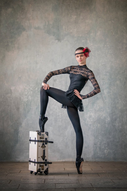 年轻的芭蕾舞女演员穿着黑色西装和黑色尖头鞋，在一堵纹理分明的墙的背景下跳舞，带着一个白色手提箱，摆出一种优雅的姿势图片下载