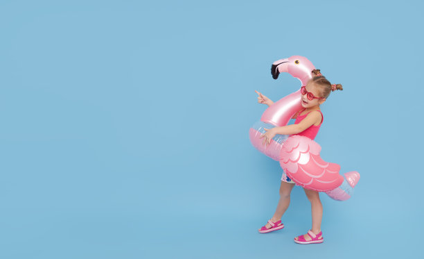 一个快乐的女孩穿着泳衣，戴着火烈鸟充气戒指要去度暑假了。向左边伸出手指。蓝色背景图片下载