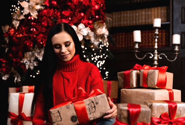 正女孩的全身照片，帽子拿着大礼盒在圣诞之夜享受新年传统坐在地板地毯上。图片下载