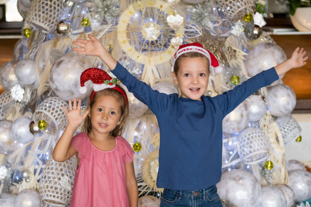 两个快乐的孩子兄妹站在圣诞树旁挥手致意的人像照片。圣诞快乐，节日快乐图片下载