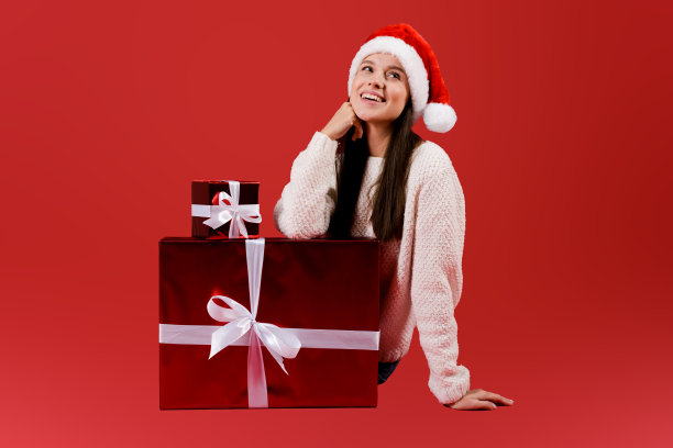 年轻女子坐在用白色蝴蝶结包着的红色礼盒旁边，戴着红色的圣诞老人帽，在红色的演播室背景下与世隔绝图片下载
