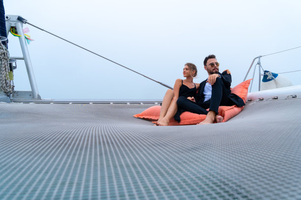 豪华放松的夫妇旅行者在漂亮的衣服和套房坐在豆袋在部分游艇与海和白色的天空的背景。商务旅行概念。图片下载