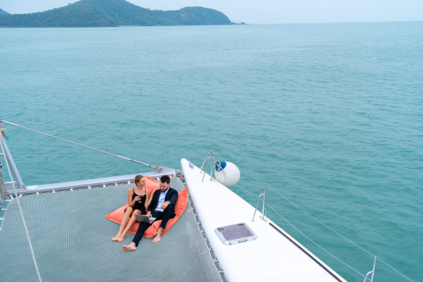 豪华放松的夫妇旅行者在漂亮的衣服和套房坐在豆袋和看电脑的一部分，在海上和白色的天空的背景游艇。商务旅行概念。图片下载