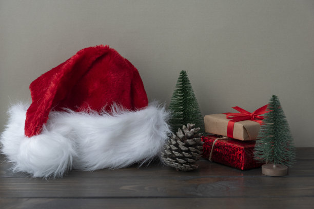 毛茸茸的红白圣诞帽，包装好的礼盒，松果和玩具松树放在木桌上。图片下载