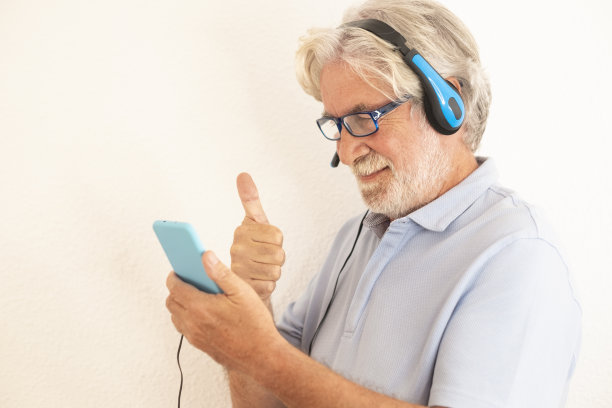 白发老人戴着耳机站在白色背景下看他的智能手机与家人或朋友视频通话。老年人享受新技术图片下载