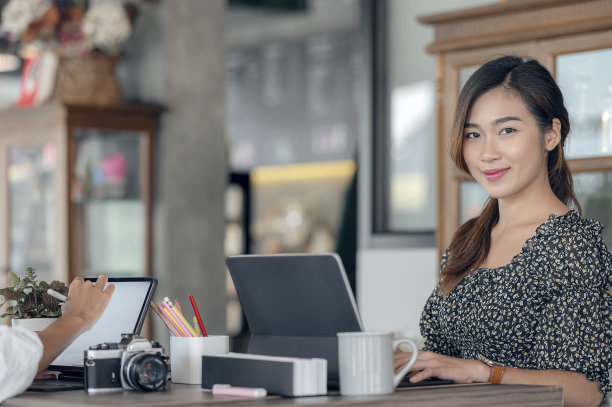 年轻美丽的亚洲女人的肖像微笑，看着镜头，而坐在她的办公桌。图片下载