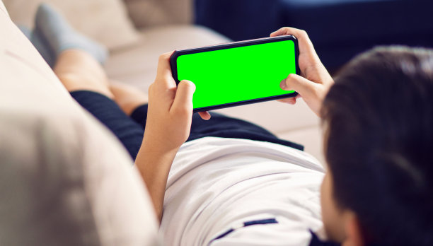坐在沙发上的孩子手持绿色屏幕触摸屏智能手机。浏览社交网络，阅读新闻或使用应用程序或玩游戏。社会成瘾概念。色度键。图片下载