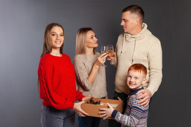 这个幸福的家庭微笑着，在灰色的背景下在工作室里庆祝节日。妈妈和爸爸拿着玻璃杯里的香槟，孩子女孩和男孩拿着一盒甜甜的蛋糕。图片下载