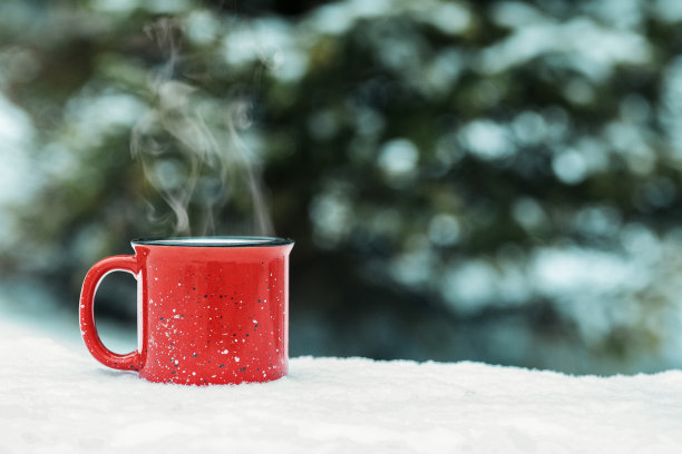 红色的杯子里放着冬天的热饮(热葡萄酒、可可、咖啡、茶)，背景是冬天的森林和积雪。冬天心情舒畅。图片下载