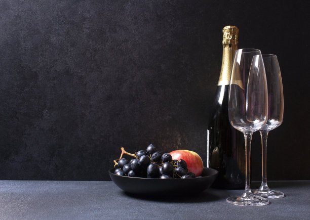 一瓶香槟，两个杯子，一盘水果。在黑色的背景上。黑暗的照片。图片下载