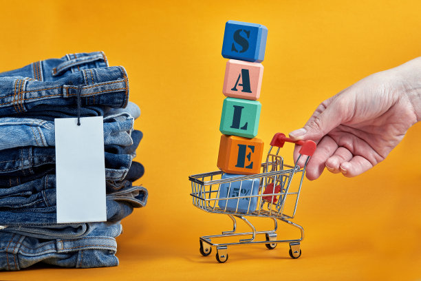 黄色背景上有白色空白标签的一堆蓝色牛仔裤。购物车与多色立方体。销售文字写在立方体上。销售consept图片下载