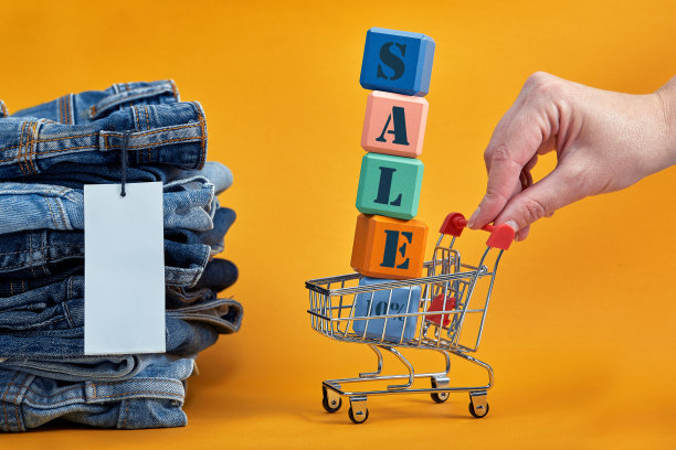 黄色背景上有白色空白标签的一堆蓝色牛仔裤。购物车与多色立方体。销售文字写在立方体上。销售consept图片下载