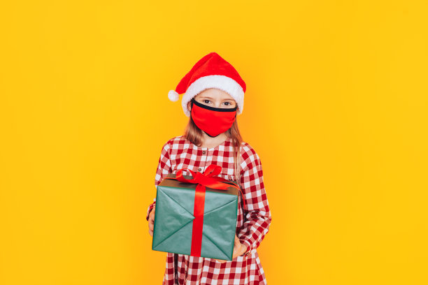 拿着装有礼物的大盒子的孩子。她头上戴着圣诞老人帽，脸上戴着医用口罩。圣诞节的心情。大流行期间，一张完美的圣诞广告照片图片下载