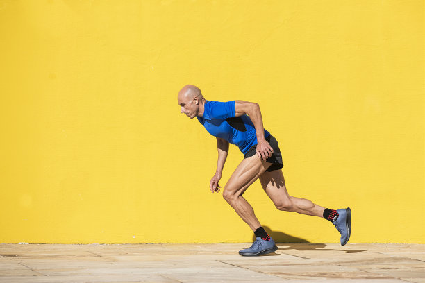 剃着光头、穿着运动服的成年男子开始在一堵黄色墙前的街道上冲刺图片下载