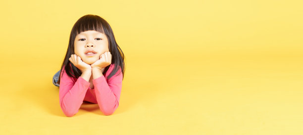 可爱的亚洲小女孩躺在地板上，双手放在下巴孤立在黄色的背景。宽裁剪，长水平横幅，横幅为网站标题概念。图片下载