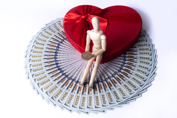 一个有象征意义的人的木制小雕像，手里拿着一美元硬币，坐在礼品盒上，盒子上有一个心形的人的象征，上面有100美元的钞票图片下载