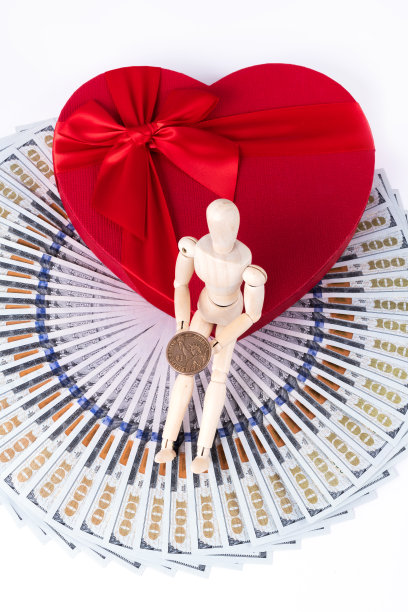 一个有象征意义的人的木制小雕像，手里拿着一美元硬币，坐在礼品盒上，盒子上有一个心形的人的象征，上面有100美元的钞票图片下载