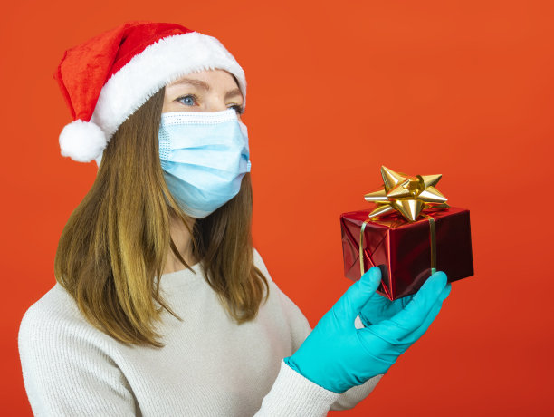 年轻女子用防护面具和防护手套在红色圣诞背景下给圣诞礼物，圣诞节与社会距离素材图片