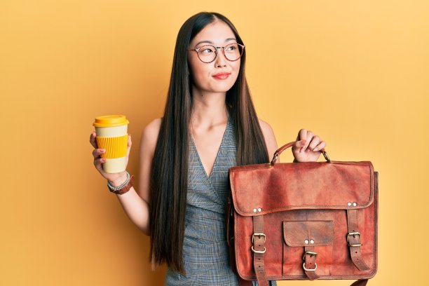 年轻的中国女人拿着皮包，喝着带走的咖啡微笑着看向一边，盯着远方思考。图片下载