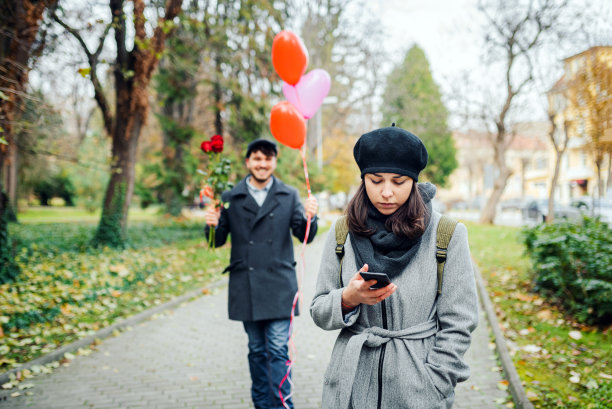 年轻紧张的女人检查她的手机上的时间为她的约会和年轻的男人惊喜她与玫瑰花束和气球股票照片。情人节的概念。图片下载