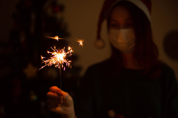 年轻女子戴着面具，手持烟花，站在圣诞树旁庆祝。图片下载