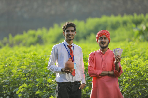 年轻的印度银行家或农学家和农民展示钱和粘土小猪银行在田间图片下载