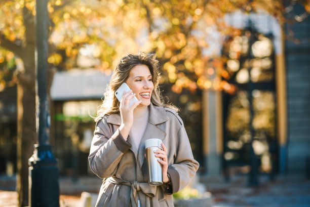 一个美丽的微笑的年轻白人妇女的肖像在电话中交谈。秋天的户外/城市街道照片。图片下载