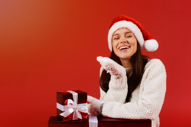美丽的年轻女子在红色圣诞老人帽子和白色针织手套坐在红色礼盒和吹空气吻孤立在红色背景图片下载