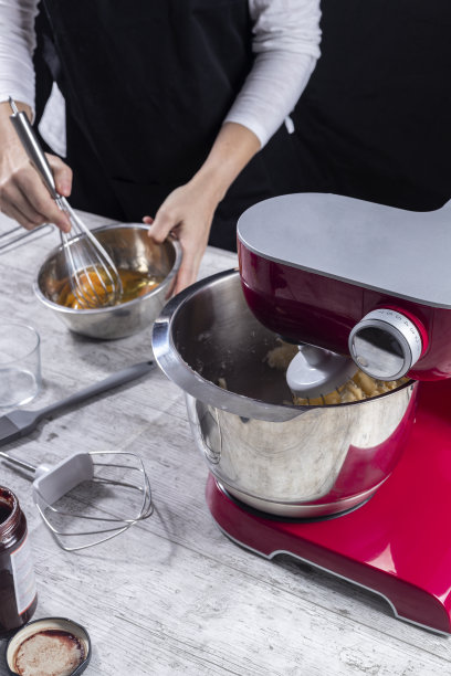 搅拌器工作和后面的女人打鸡蛋用她的手。烹饪和糕点的概念在家里，博客，课程，社交网络和小型企业。垂直照片和选择焦点。图片下载
