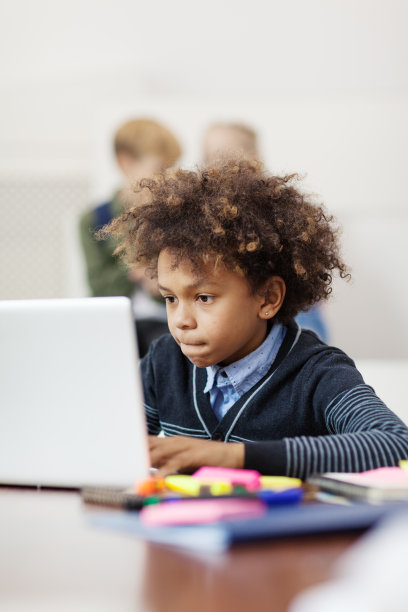 混血卷发男孩坐在课桌上用笔记本电脑学习，脸上带着沉思的表情图片下载