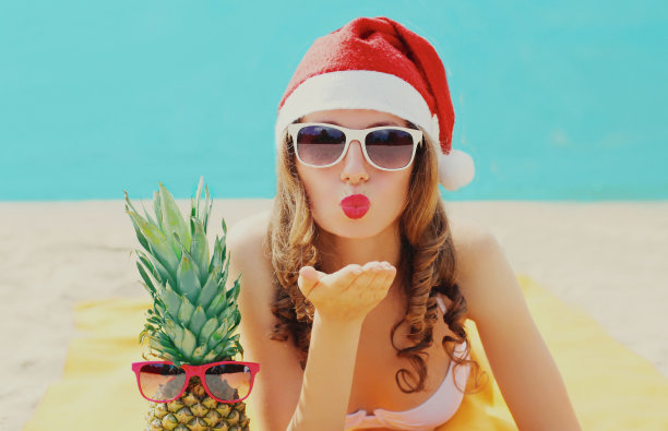 圣诞节的年轻女子吹嘴唇送甜蜜的空气吻在红色圣诞老人的帽子与菠萝躺在一个海滩上蓝色的海洋背景图片下载