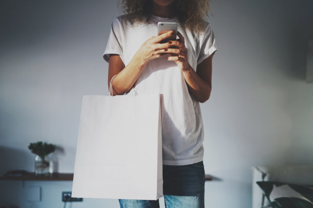 年轻的潮女拿着一个空白的白纸包装在她的手与标志和设计的空白空间。裁剪的照片，一个女孩写一条短信在现代手机上。图片下载