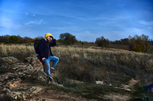 一个穿着牛仔裤、羊毛帽和背包的年轻人的侧面全长肖像坐在陡峭的岩石上，看向远方的手超过了头。天空和陡峭的背景。图片下载