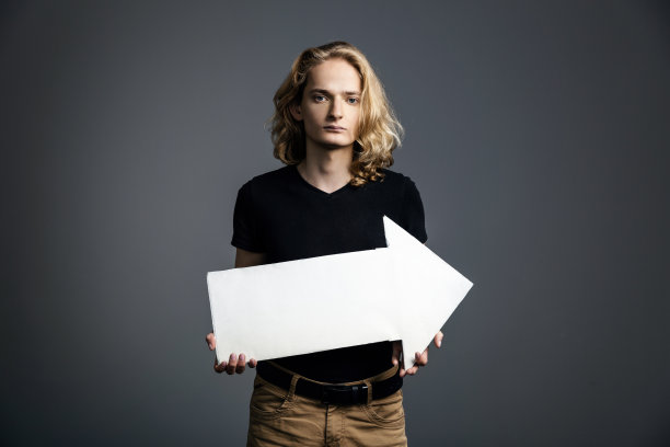 一个英俊的年轻人，有着长长的金发和一张悲伤的脸，在灰色的背景上举着一个白色的箭头指向右边。图片下载