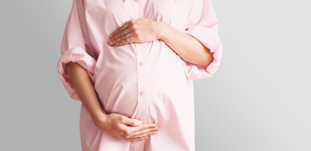 白色背景下，身着粉色衬衫裙的美丽孕妇手放在腹部的特写。图片下载