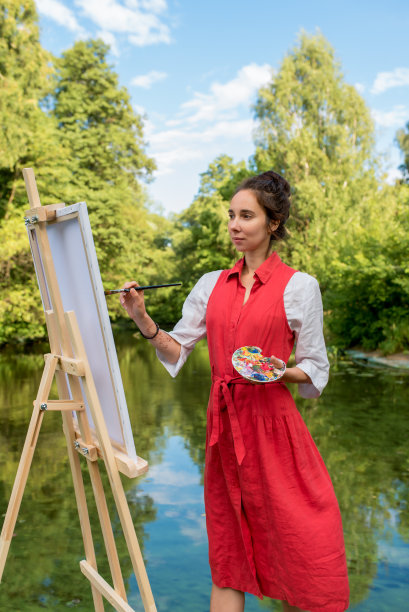 女艺术家夏天在河边的公园里画画，在湖边和池塘边创作风景背景。画笔和调色板。红色连衣裙和白色衬衫。在森林的水。图片下载