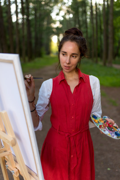 美眉艺术家在夏日森林中，特写、画图、山水背景创作创意。站在画架上作画。时尚的红裙白衬衫，调色板和油漆刷。图片下载