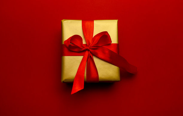 节日礼物。盒子在一个金色的礼品包装与一个红色的蝴蝶结，在一个红色的背景。圣诞快乐，新年快乐，情人节问候。给爱的人寄明信片。图片下载