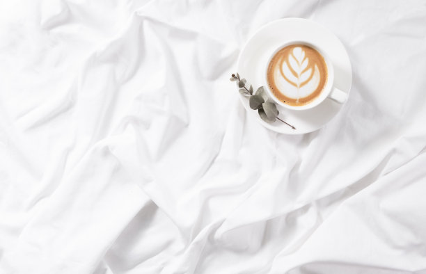 在床上喝一杯卡布奇诺咖啡。早晨平躺在白色的床上。图片下载