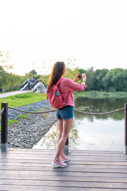 女人夏天在城市附近的湖，河池塘，照片风景智能手机，录制视频到互联网，应用智能手机摄像头。水树背景，码头，码头。休闲穿粉红色夹克。图片下载