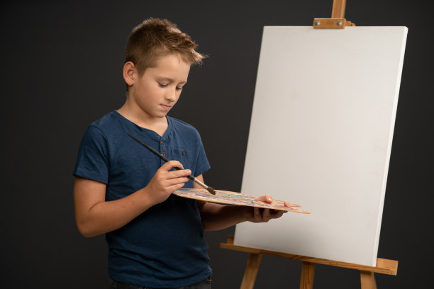 10岁的小男孩穿着蓝色t恤，拿着调色板，用画布看着画架上的相机，得到了想要的颜色。艺术学院的概念。艺术的概念图片下载